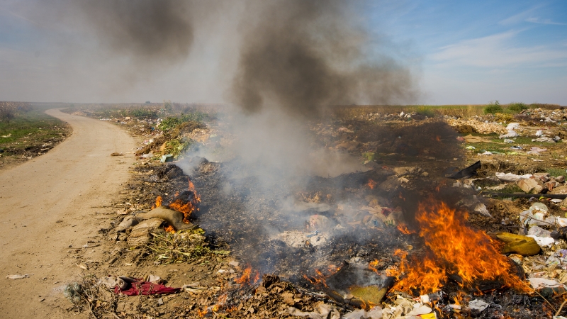 Plastic trash burns along a roadside dump, sending dark smoke into the air