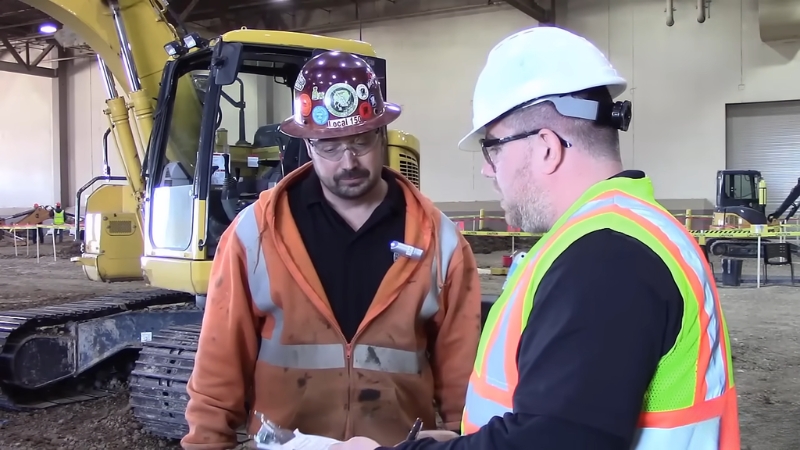 Two construction workers in hard hats review paperwork beside heavy equipment at a job site