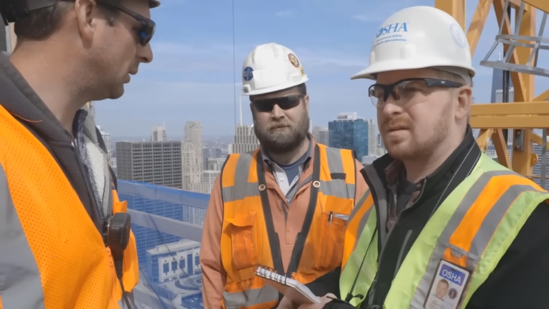 Construction workers wearing hard hats and safety vests review notes during a job site inspection