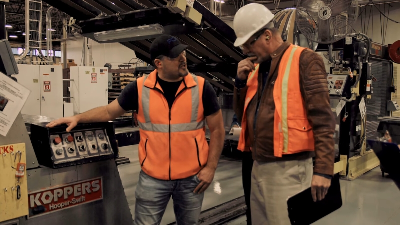 Two workers in safety vests and hard hats inspect industrial machinery inside a factory