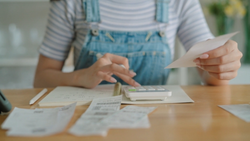 Young adult reviews bills and receipts while using a calculator at a kitchen table