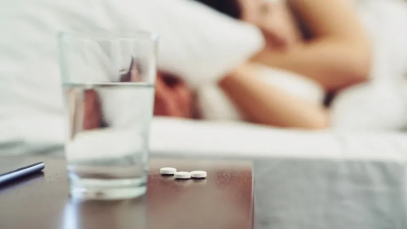 Glass of water and sleeping pills on a bedside table with a person lying in bed in the background