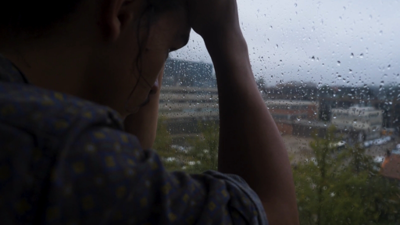 Person sits by a rain-covered window with head lowered, suggesting loneliness and distress