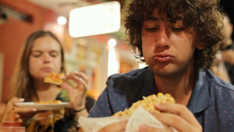 Young man eating a sandwich quickly while a woman eats pizza in the background, a common trigger for hiccups