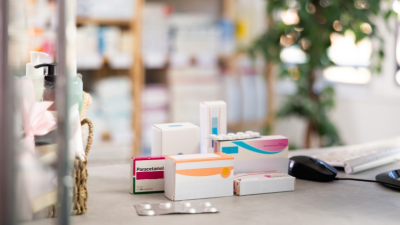 Boxes of over-the-counter medicines on a pharmacy counter with a computer and shelves in the background