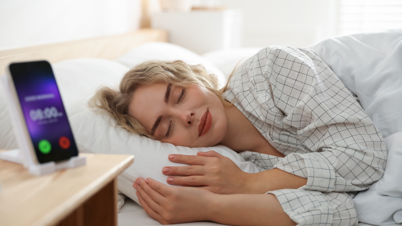 Woman sleeps peacefully beside a phone alarm clock on a bedside table