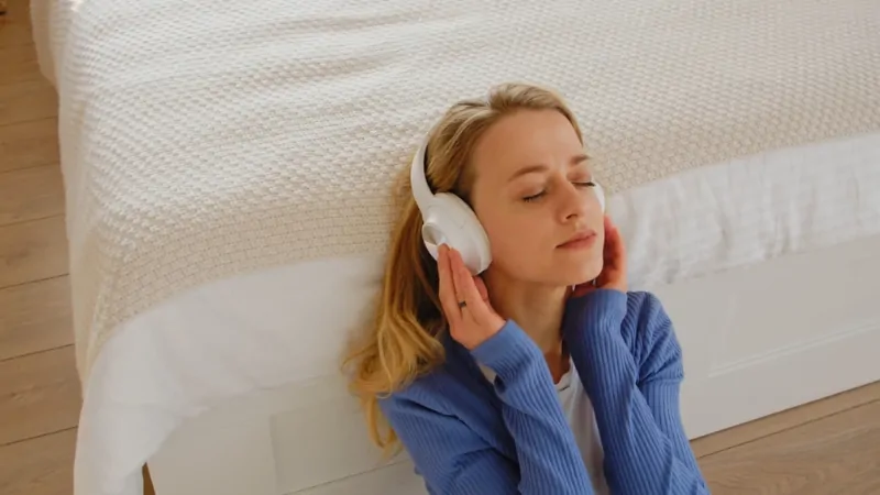 Woman sitting on the floor by her bed with headphones, listening to music with eyes closed