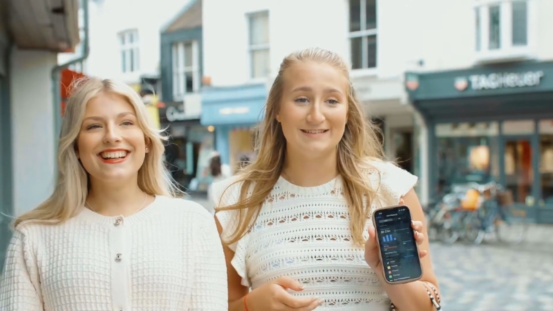 Two young women walk outside while one shows phone screen time stats on her device