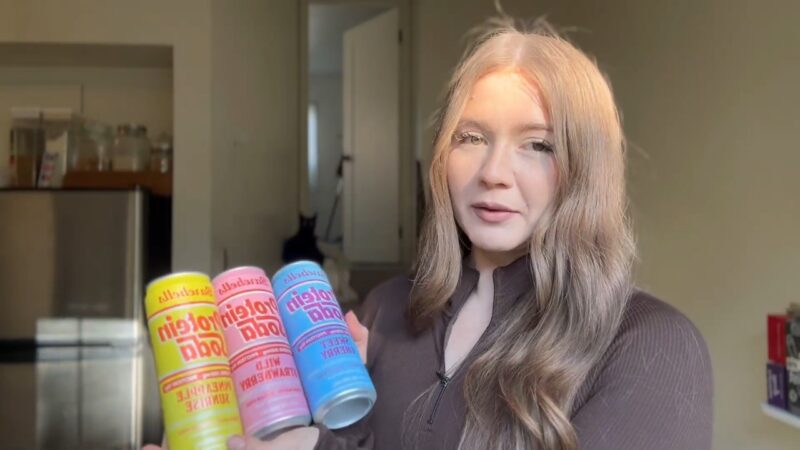 Woman holds several cans of protein soda in a home kitchen