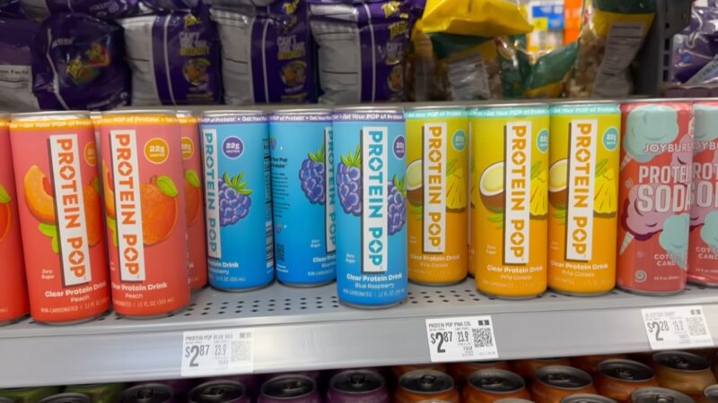 Colorful cans of protein soda lined up on a store shelf with price tags below