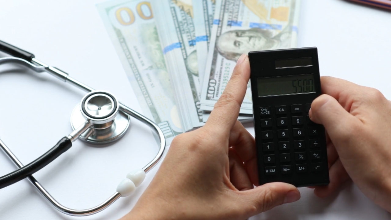 A person uses a calculator beside cash and a stethoscope to estimate healthcare costs