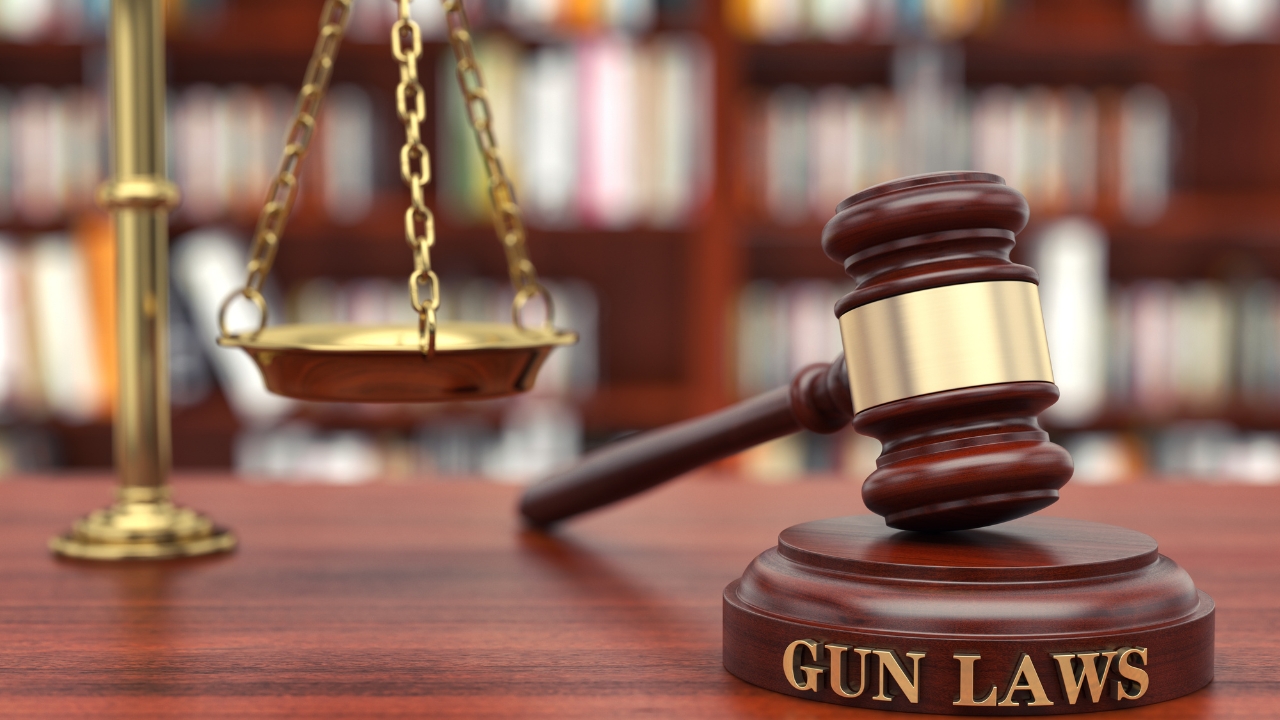 A gavel and scales sit on a desk with a gun laws sign in front