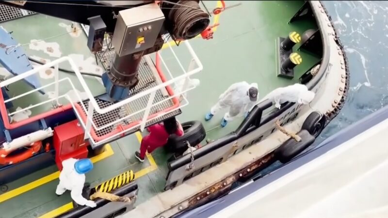 Crew members in protective suits stand on a small boat beside the MV Hondius during the health response
