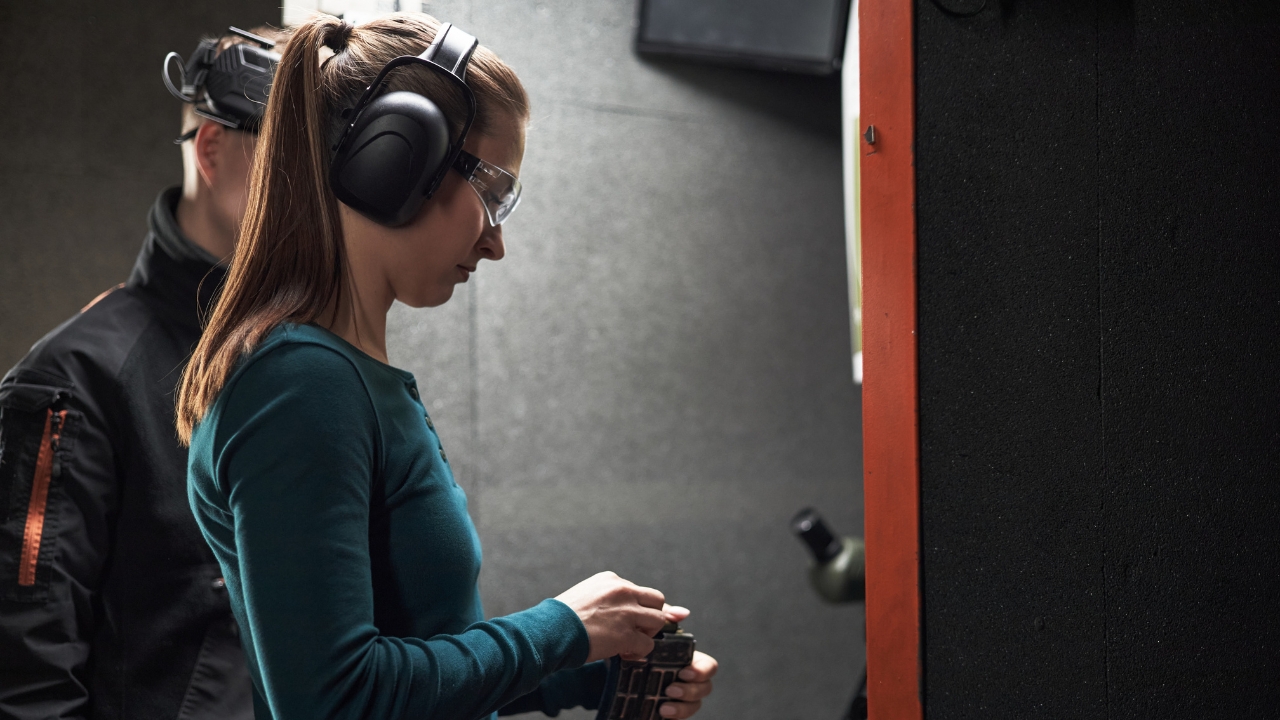 A woman wears safety gear while handling a firearm magazine at an indoor range