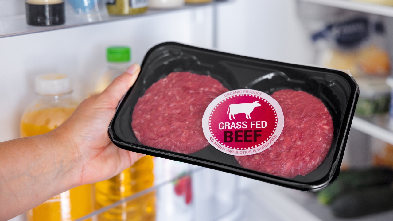A hand holds a grass-fed beef package in front of an open refrigerator