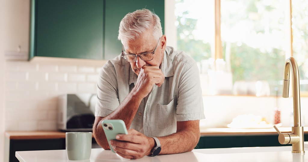 Senior man checking Social Security and inflation news on his smartphone while sitting at a kitchen table