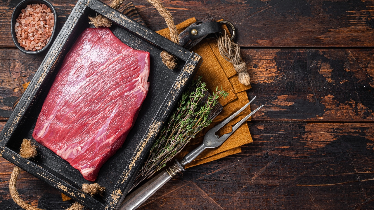 Raw grass-fed beef sits on a wooden table with herbs and salt