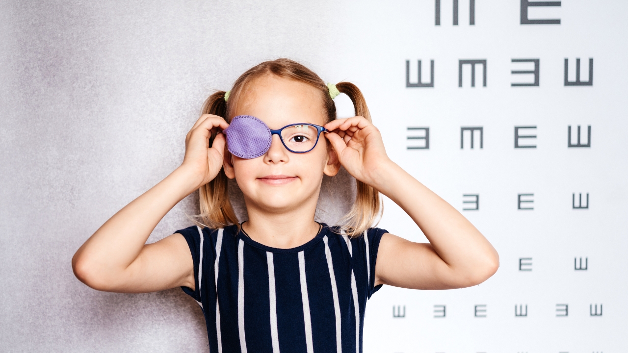Girl with an eye patch adjusts her glasses in front of an eye chart