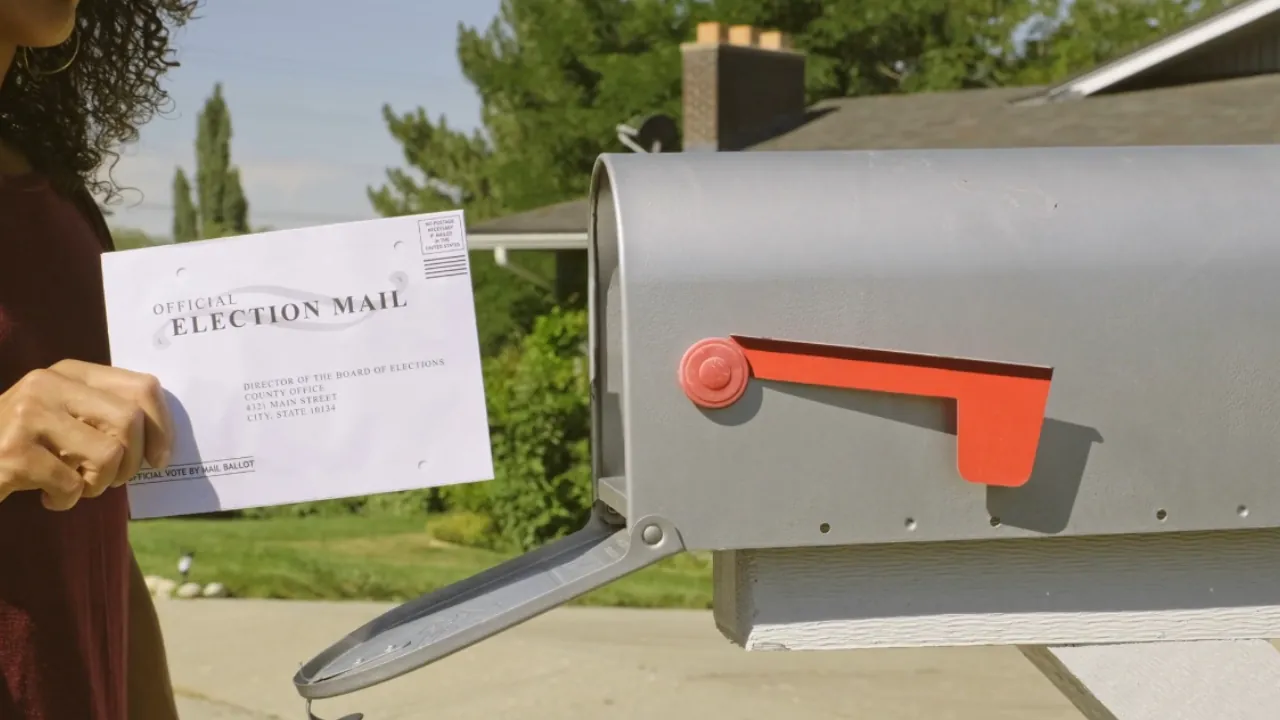A voter holds official election mail beside an open roadside mailbox