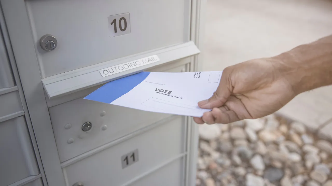 A hand places a mail ballot into an outgoing mailbox slot
