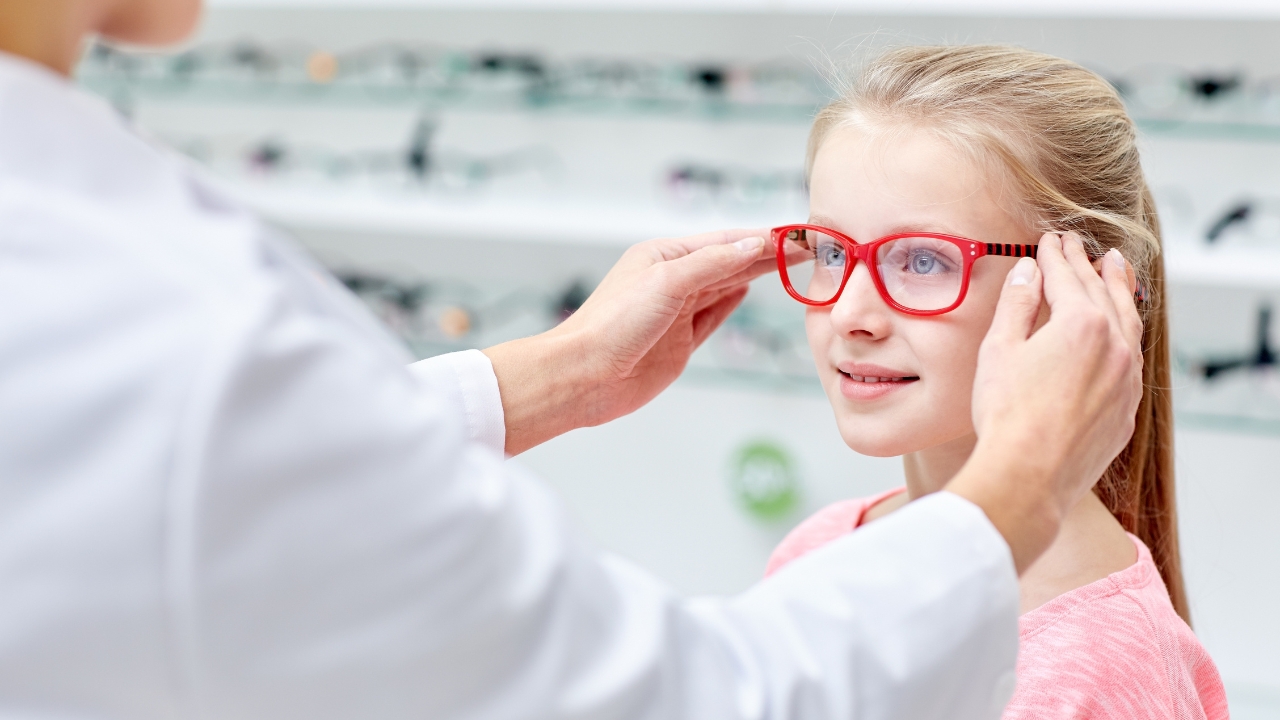 An optician fits red glasses on a young girl in an eyewear store