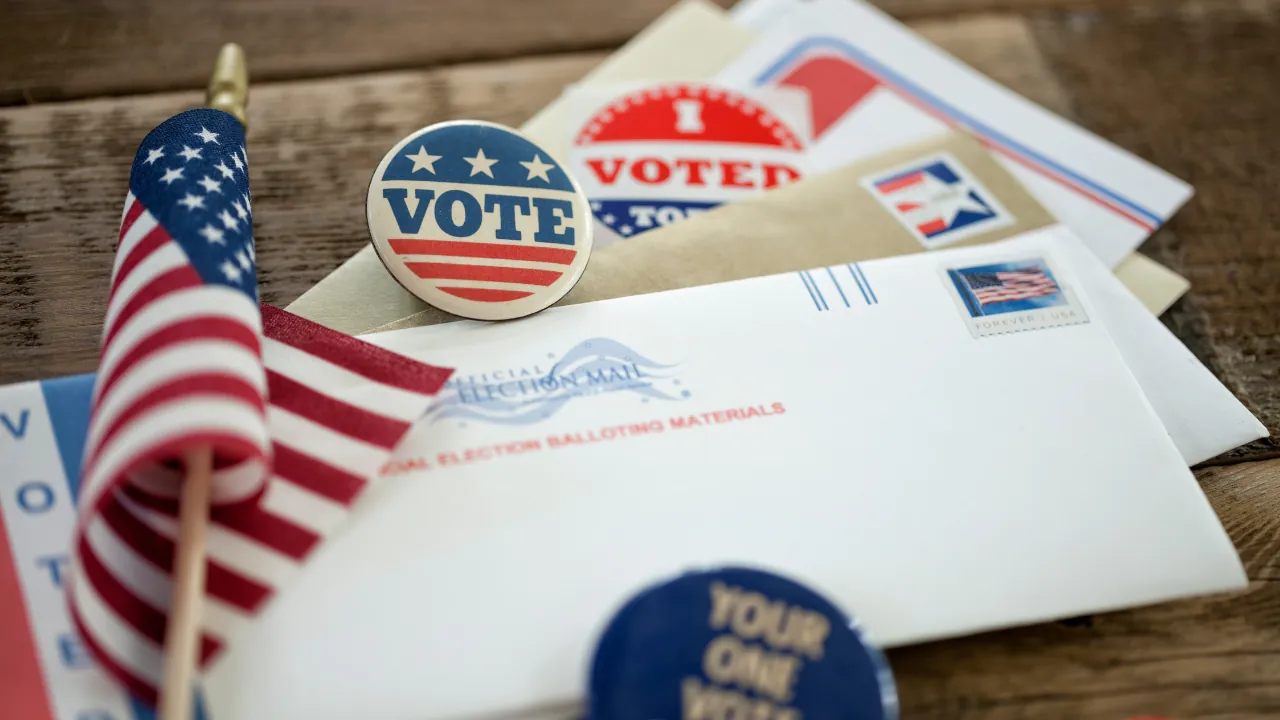 Mail-in ballot envelopes, voting stickers, and a small American flag on a wooden table