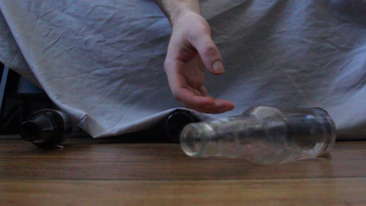 A hand hangs off a bed beside empty alcohol bottles