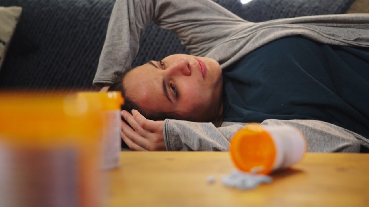 A woman lies on a couch near spilled pills and prescription bottles