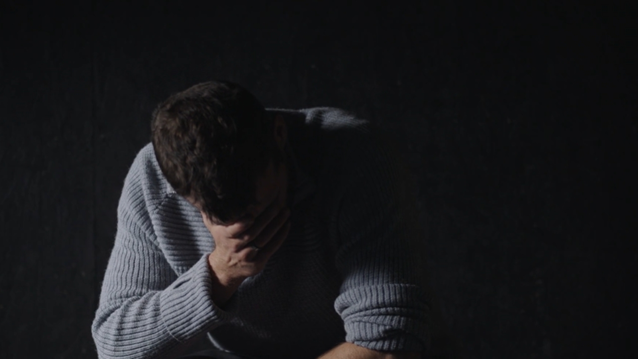 A man sits alone in a dark room with his face covered