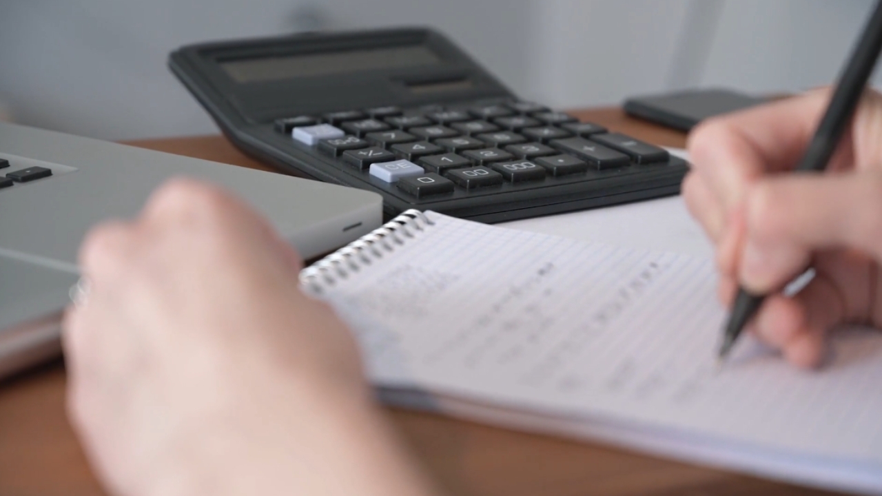 A person writes budget notes beside a calculator and laptop