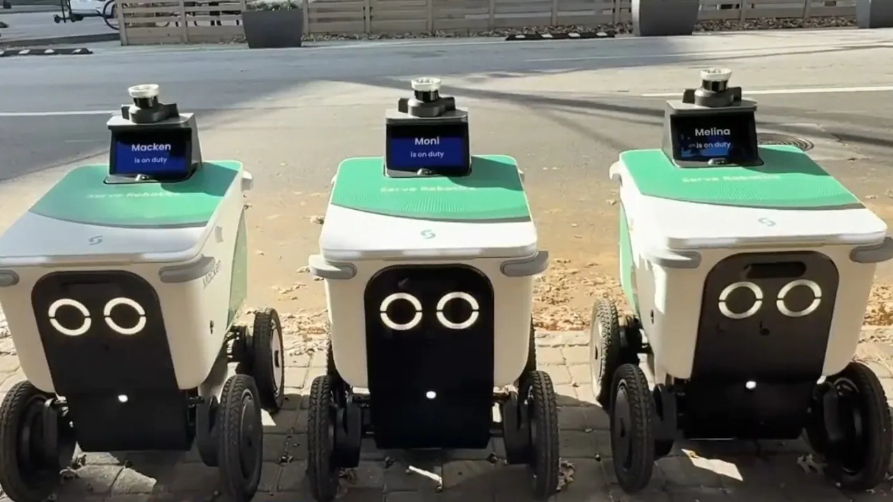 Three delivery robots stand side by side on a city sidewalk