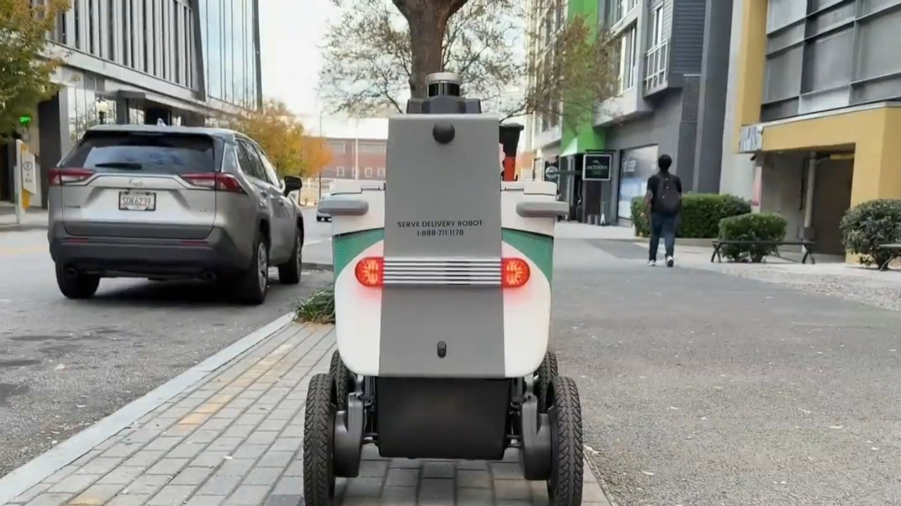 A delivery robot travels along an Atlanta sidewalk beside parked cars and pedestrians