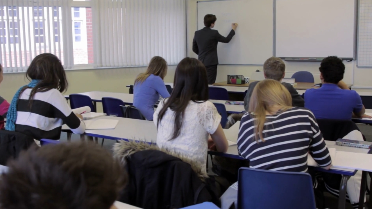Students sit in a classroom as a teacher writes on the whiteboard