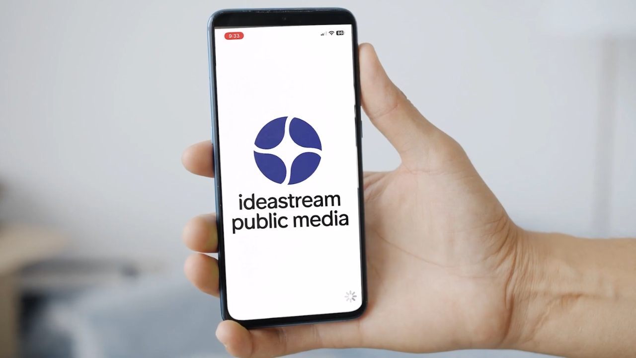 A hand holds a smartphone with the Ideastream Public Media logo on its screen