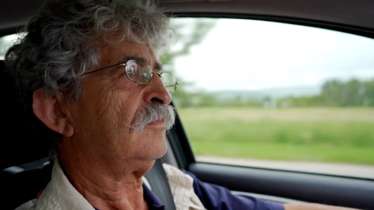 Older man with glasses drives a car along a rural road