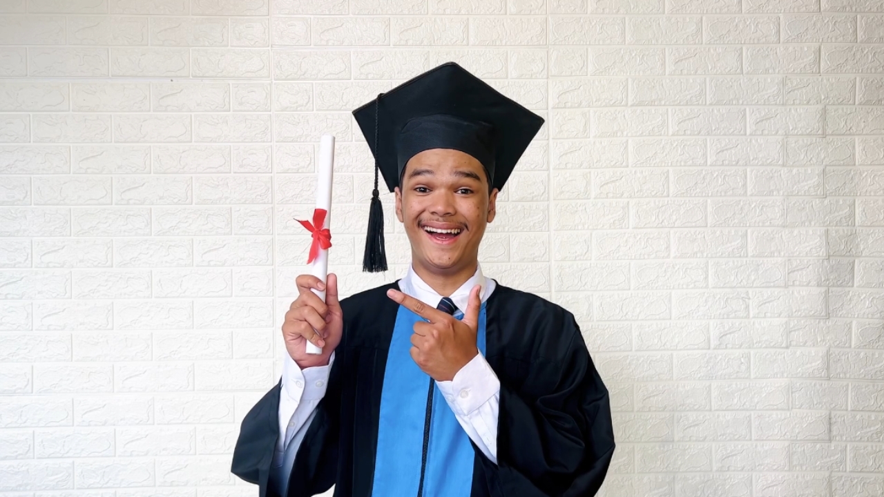 A smiling graduate points to his diploma against a white brick wall