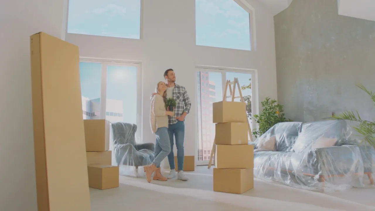 A couple stands in a bright new home with packed boxes and covered furniture