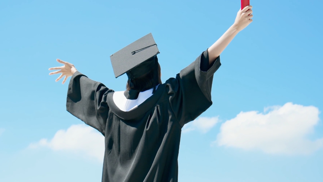 A graduate raises a diploma under a clear blue sky