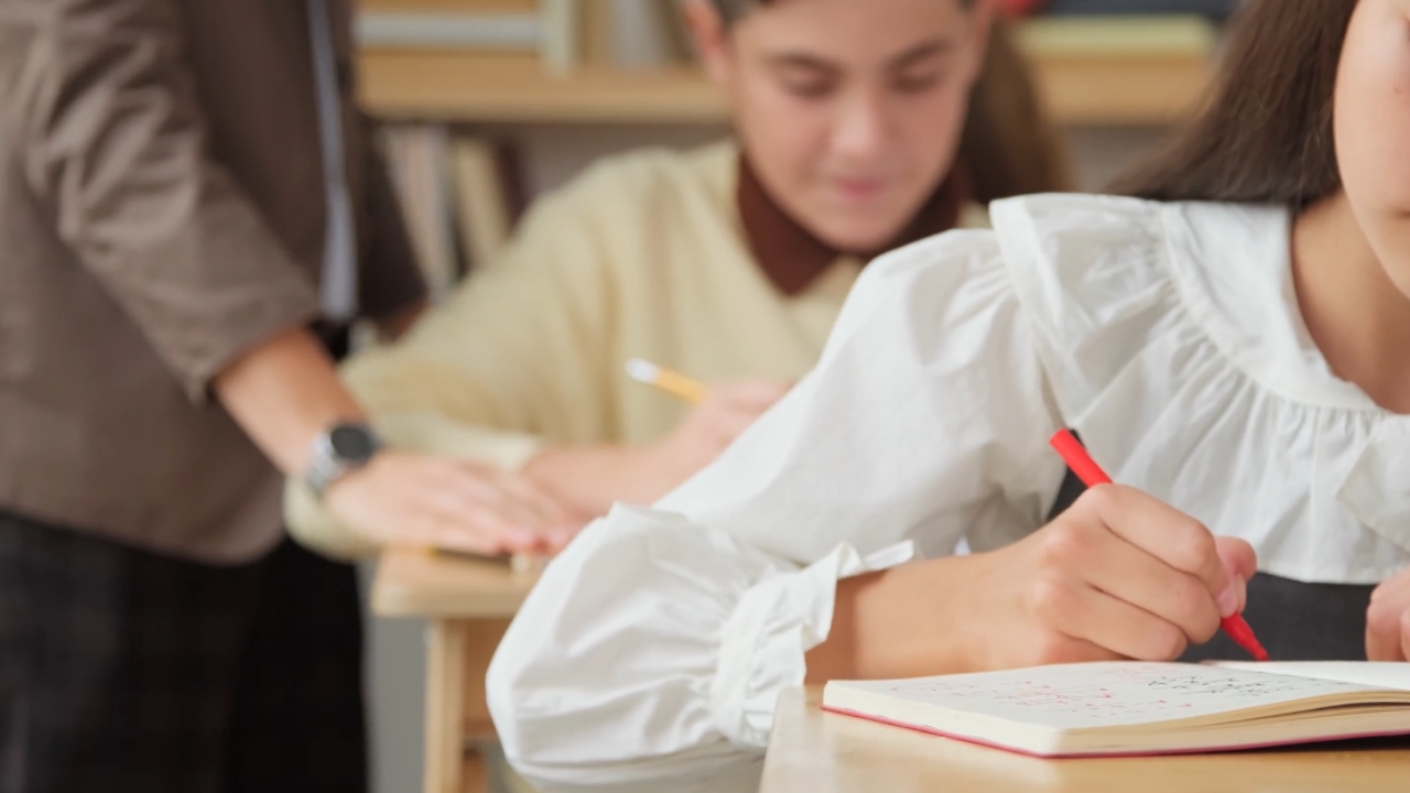 Students complete exams by hand as a teacher supervises the classroom