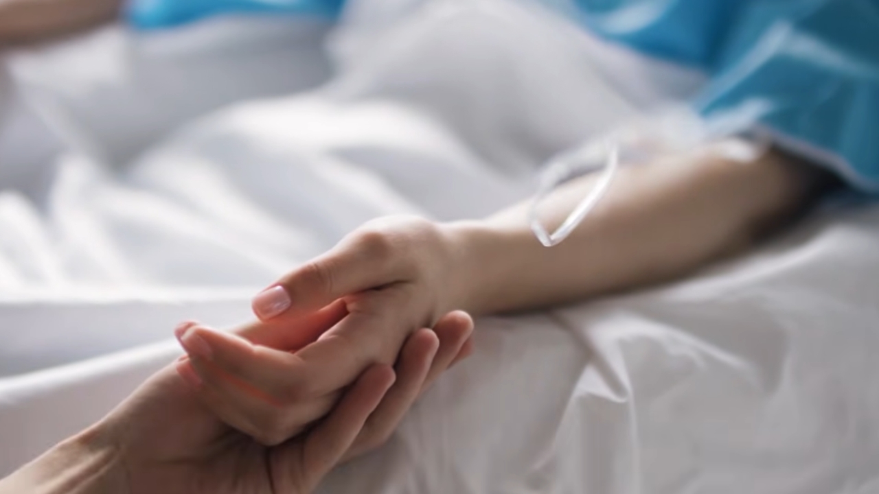 A patient with an IV rests in a hospital bed as a caregiver holds their hand