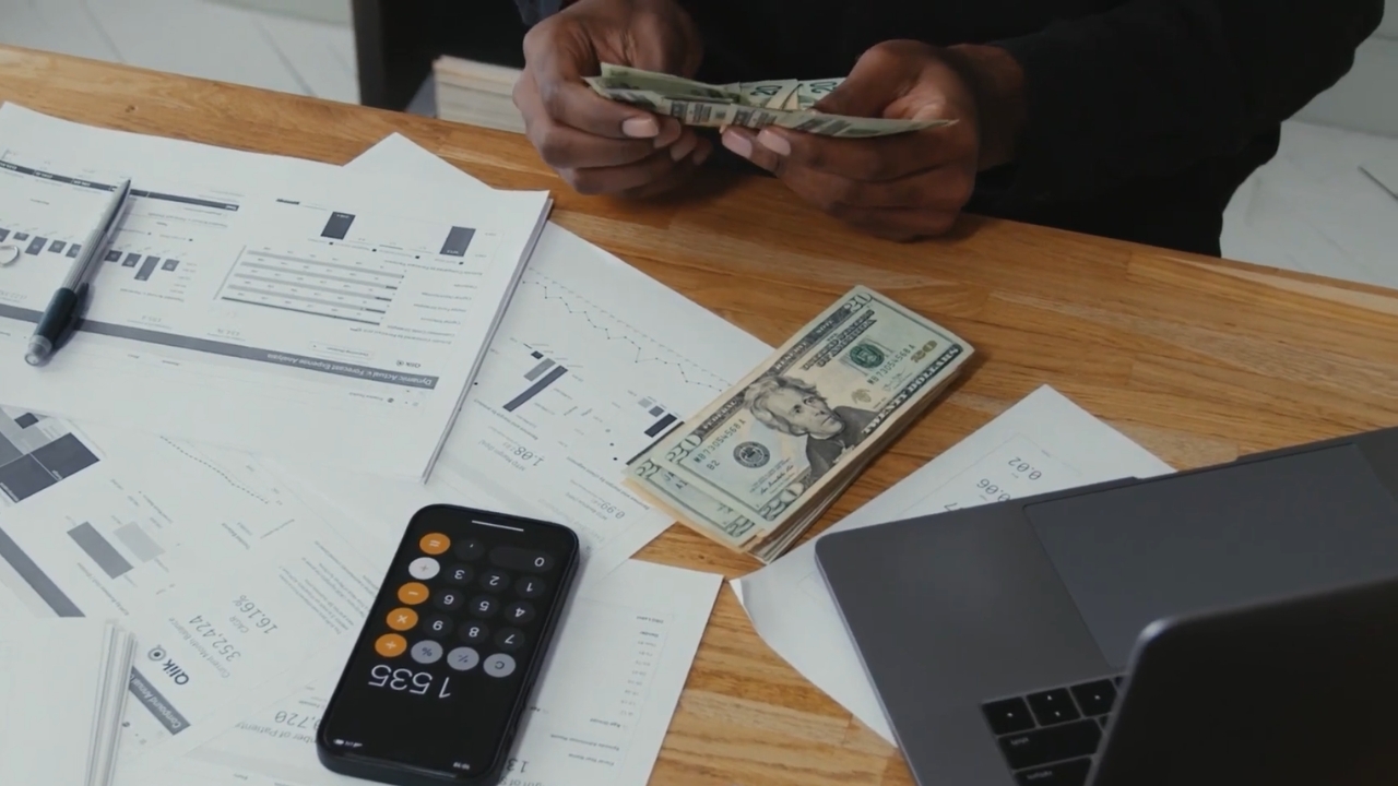 A person counts cash beside financial documents, a calculator and a laptop