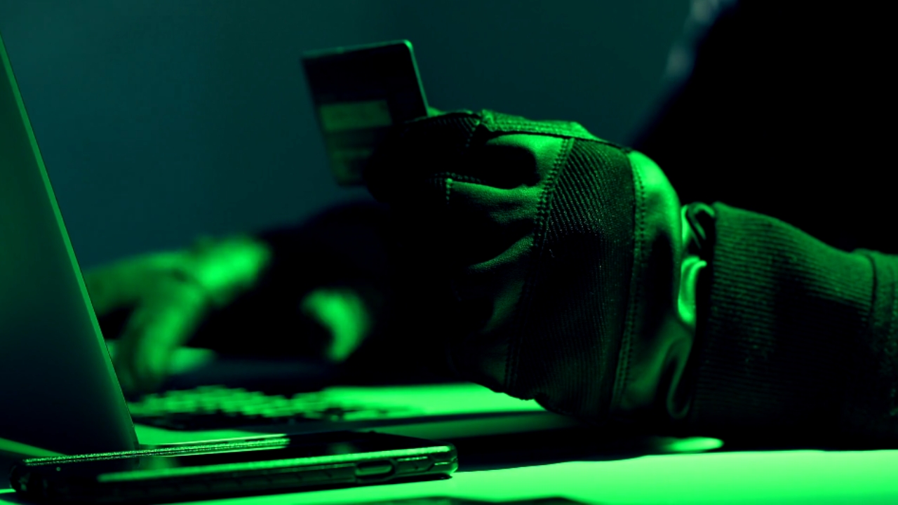 Gloved hacker holds a credit card beside a laptop under green light