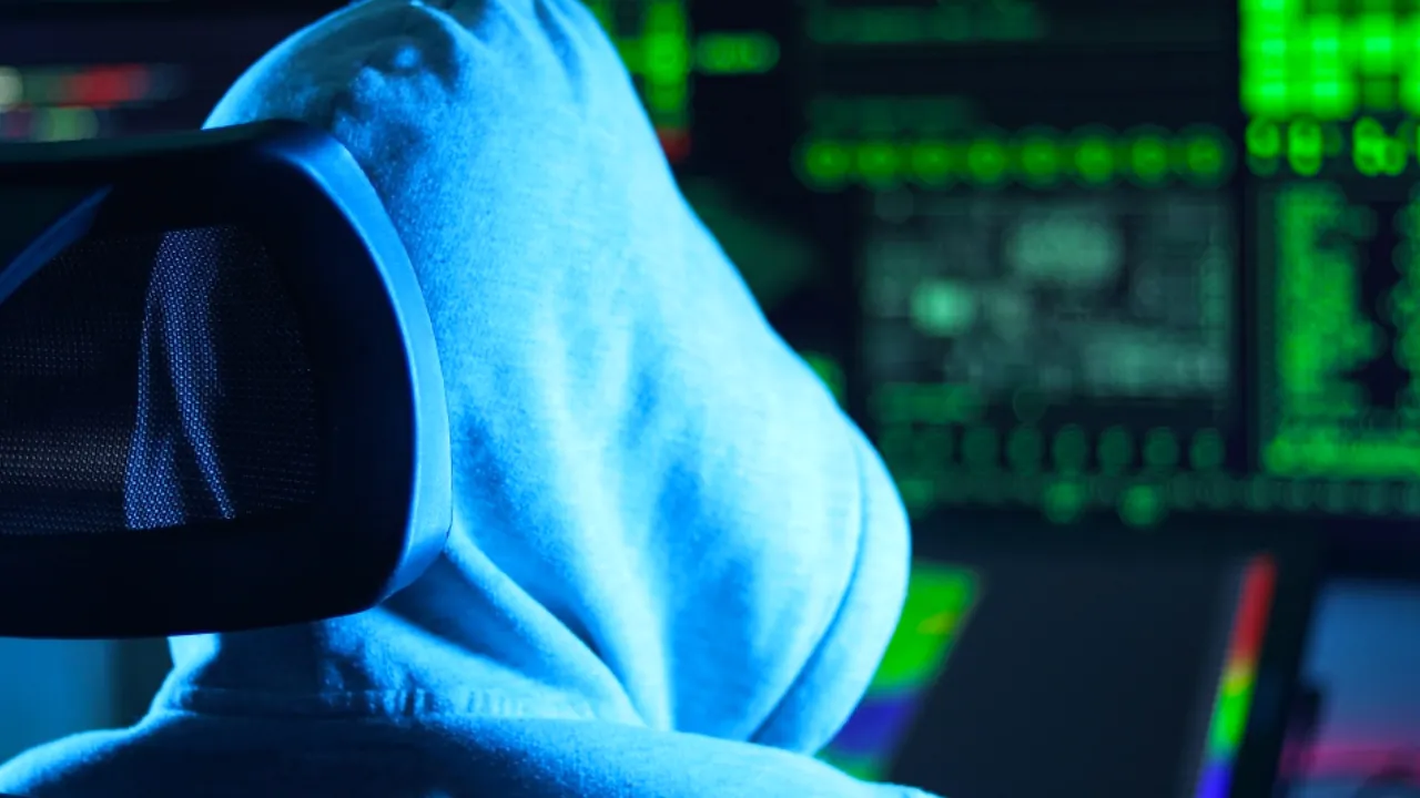 Hooded hacker sits in front of computer screens with code