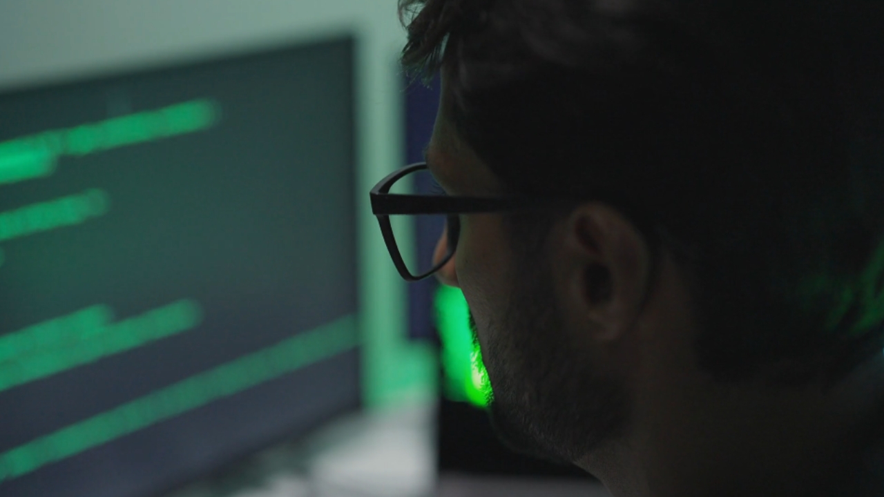 Man with glasses looks at computer screens with green code