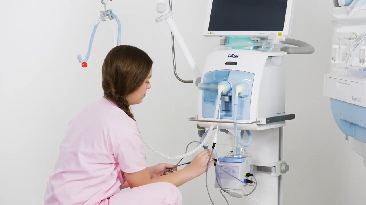 Healthcare worker checks neonatal respiratory equipment beside an infant incubator.