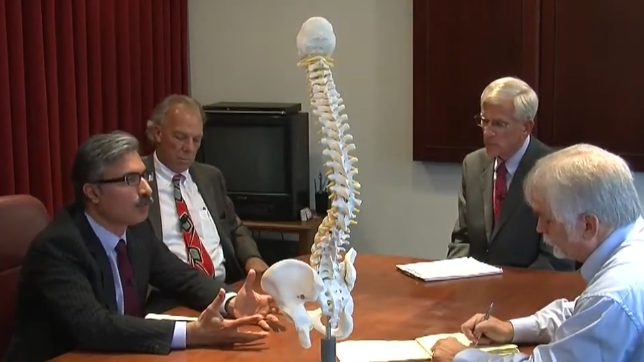 Four men discuss a spinal surgery case beside a spine model in a conference room.