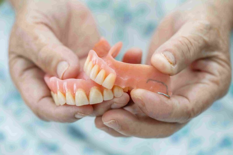 Close-up of hands holding upper and lower removable dentures with artificial teeth and pink gum-colored bases.