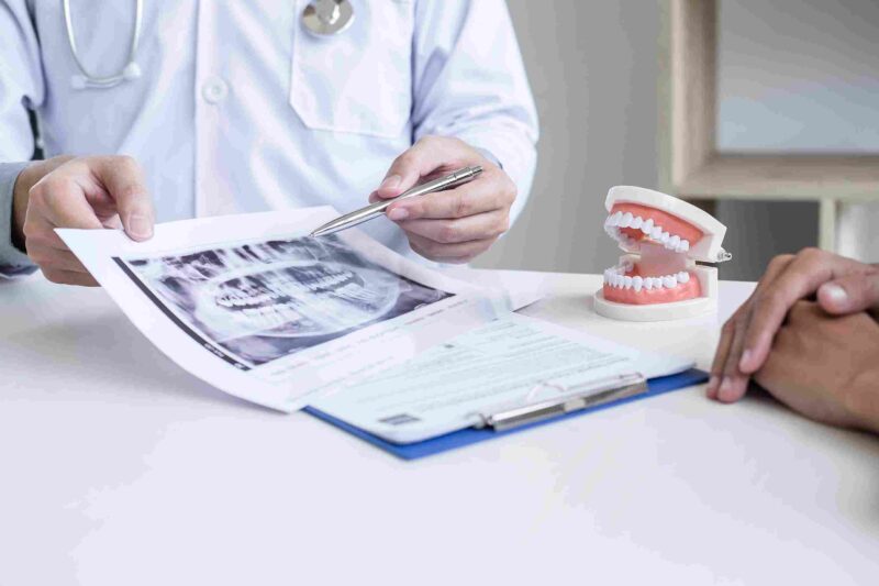 Dental professional reviewing a panoramic dental X-ray with a patient, with a model of human teeth on the desk.
