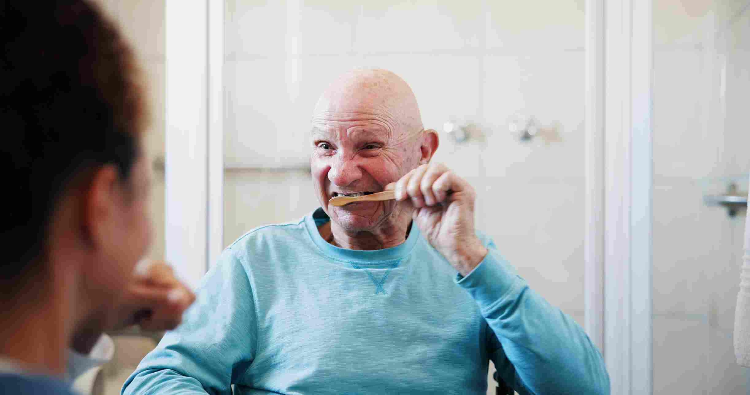 Elderly man in a blue sweater brushing his teeth while looking in a bathroom mirror.