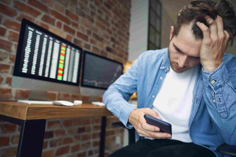 Stressed young man holding a smartphone and resting his hand on his head while sitting beside computer monitors.
